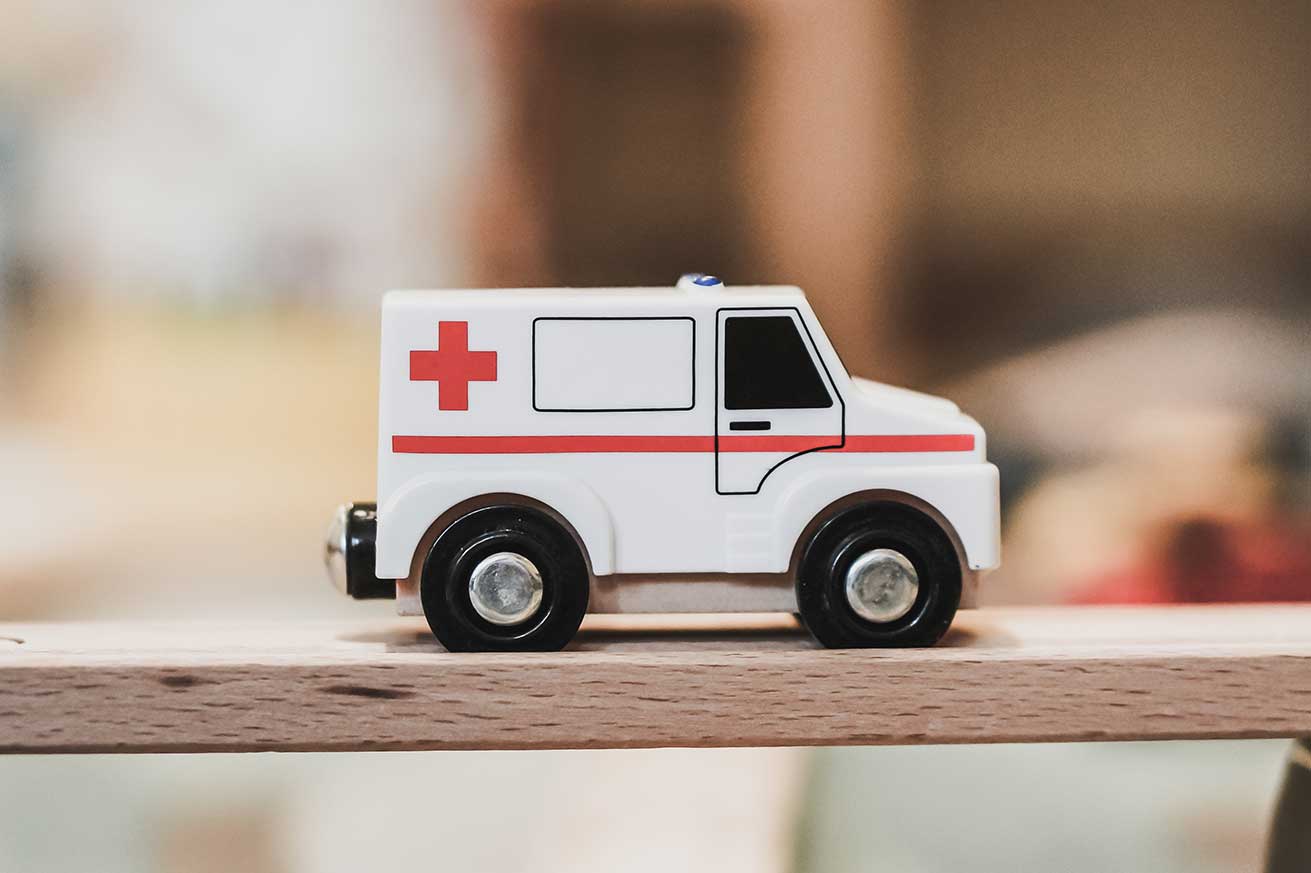 A small white toy ambulance with a red cross and red stripe sits on a wooden surface, with its black wheels visible and the background softly blurred.