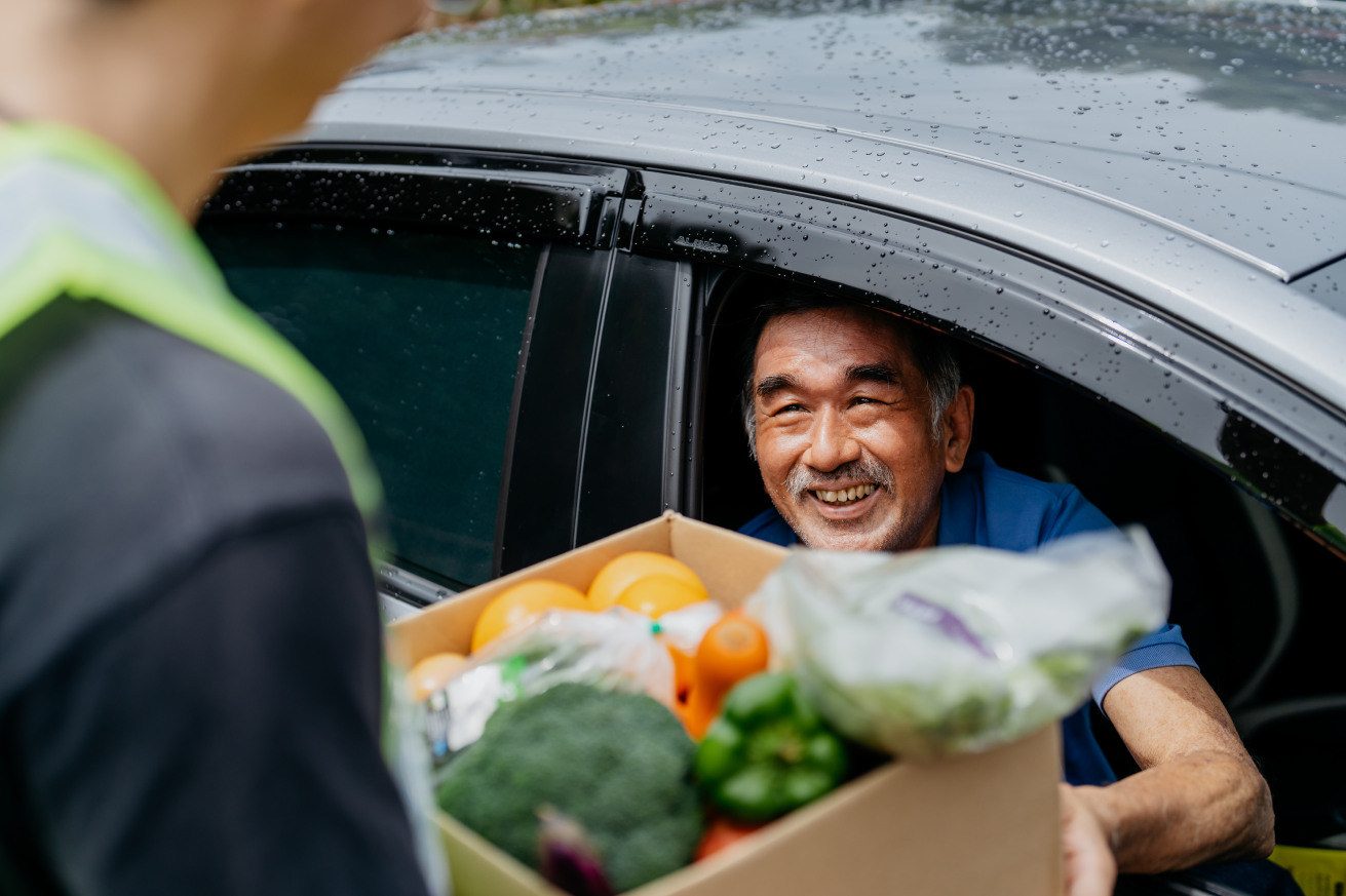 Person seated in a car receives a box of fresh groceries, including oranges, broccoli, peppers, and leafy greens, from another person standing outside the vehicle on a rainy day.