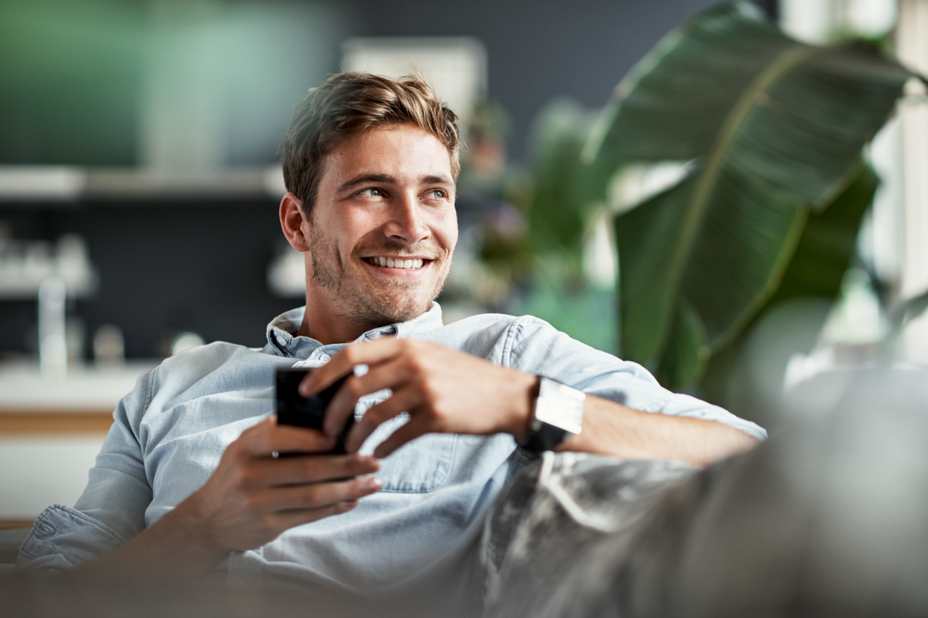 Person relaxing on a couch in a bright living room, holding a smartphone and looking to the side. A large green houseplant and softly blurred home interior are visible in the background.