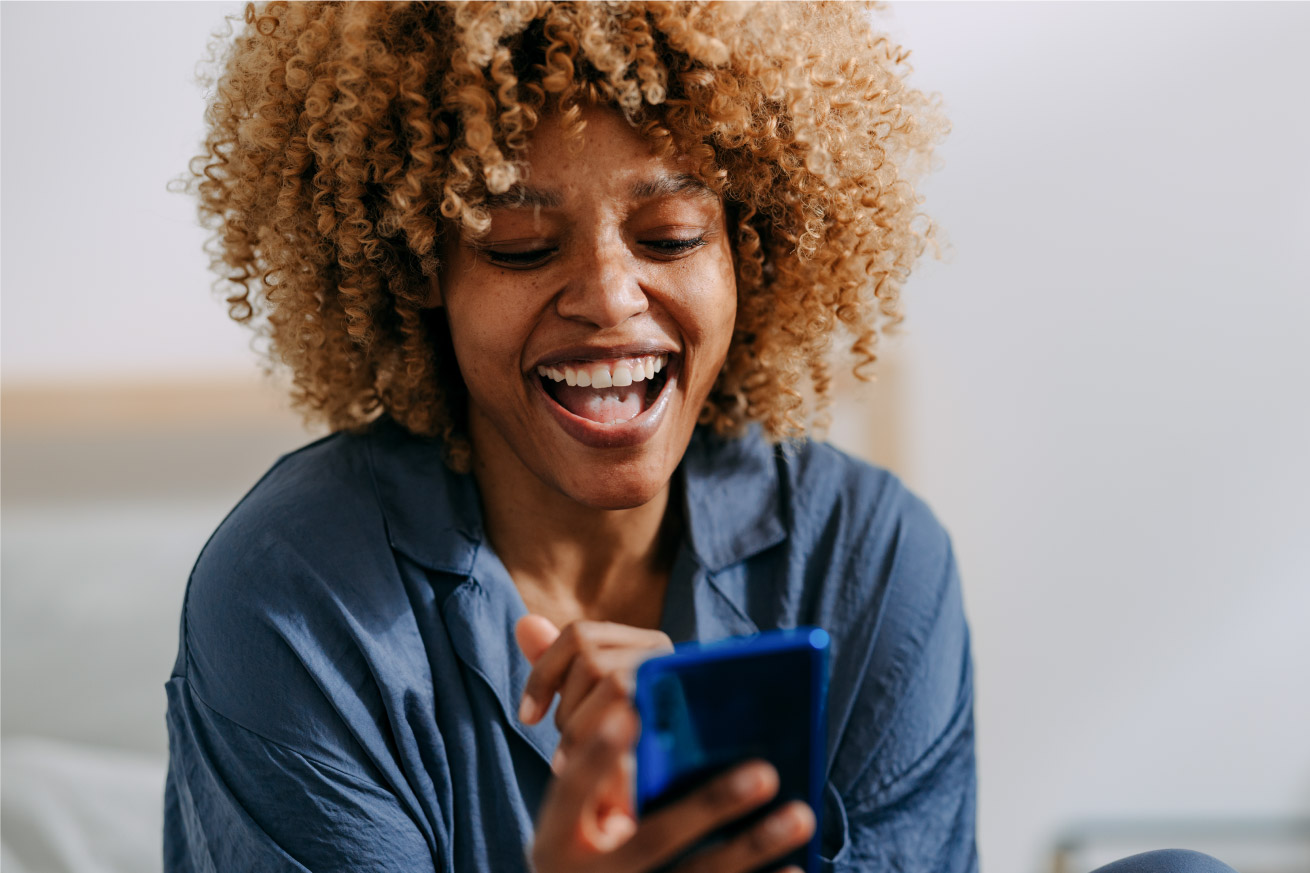 A person with curly hair is smiling and looking at a blue phone, wearing a blue shirt, in a bright indoor setting.